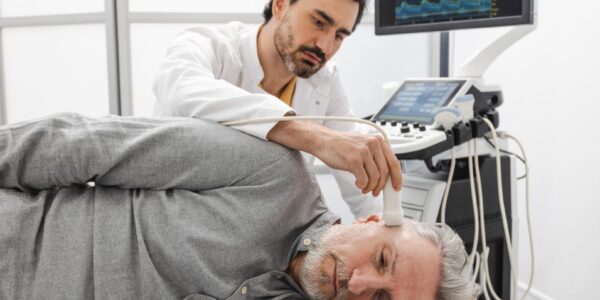 Clinician holds an ultrasound probe near a man's jaw while an imaging monitor displays scan data.